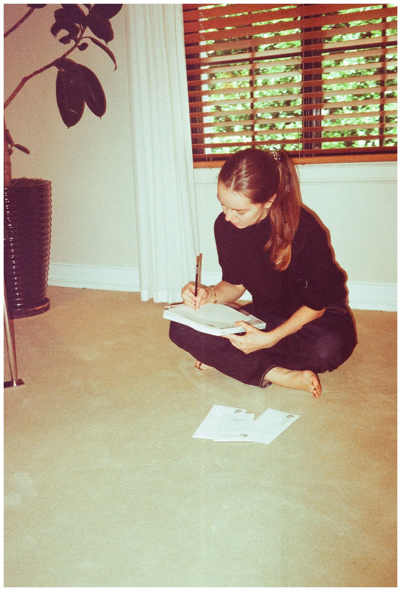 Alex Hanover sitting cross-legged on the floor of a sunlit room, writing in a notebook, with a potted plant and a shuttered window behind her.