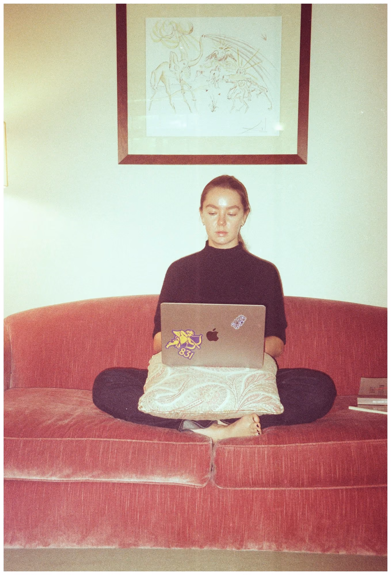 Alex Hanover sitting cross-legged on a red velvet sofa with a laptop, a framed drawing on the wall above her.